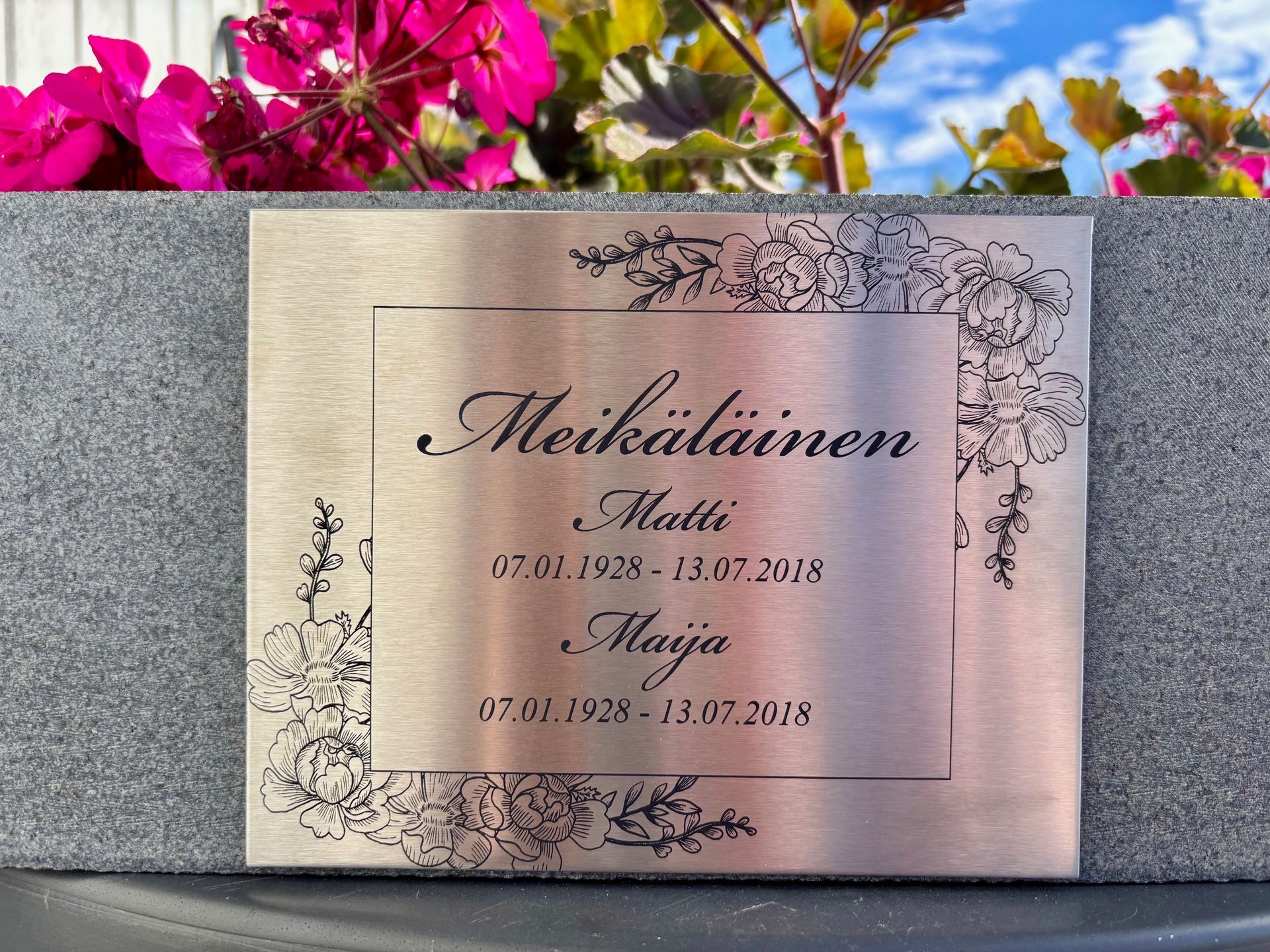 Kiveen upotettu metallinen muistolaatta, jota kehystää kukkakuvioinen reunus. 