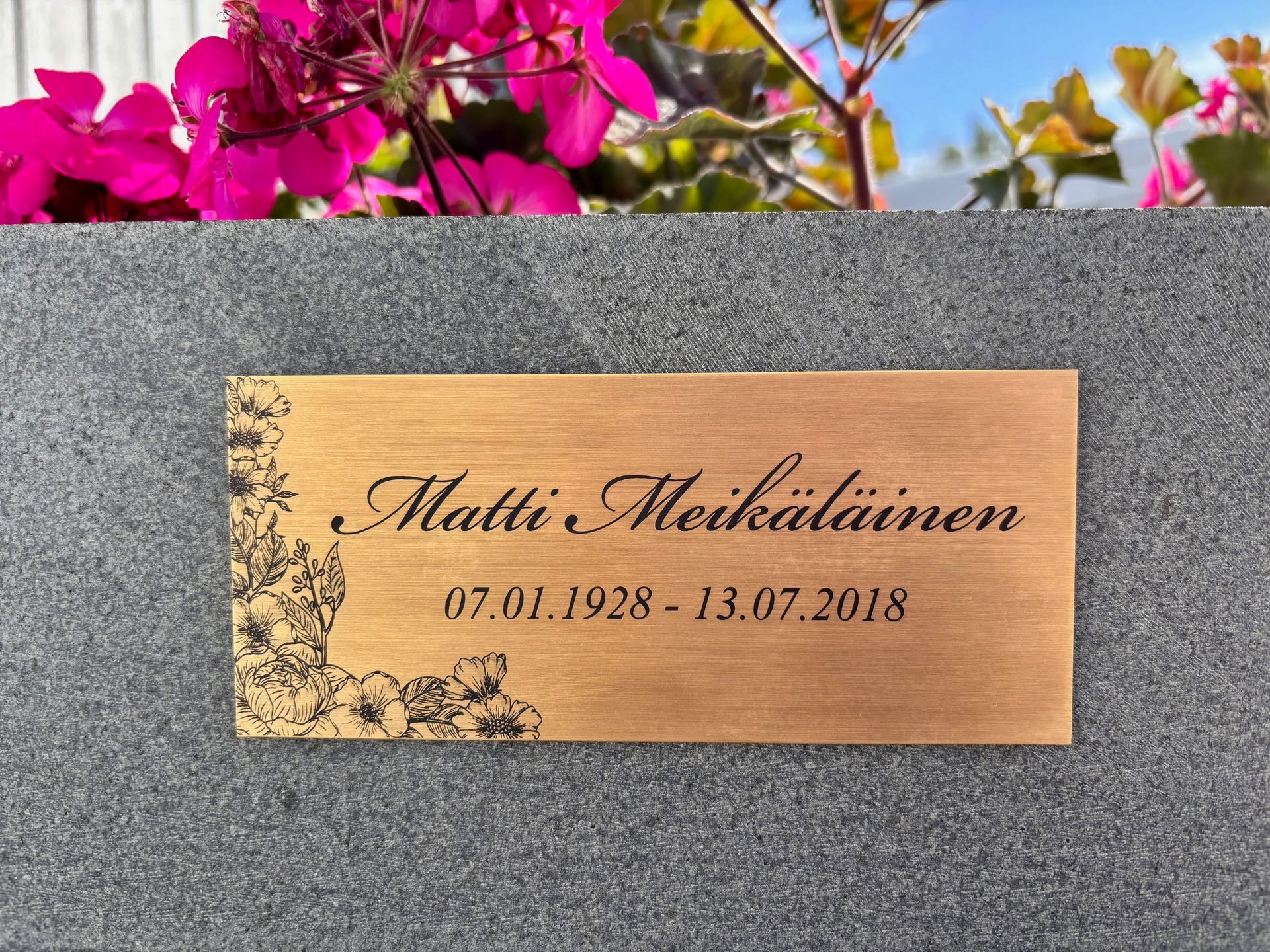 Messinkinen muistolaatta, jossa on kukkakuvioitu kaiverrus ja päivämäärät, harmaata kivitaustaa vasten.