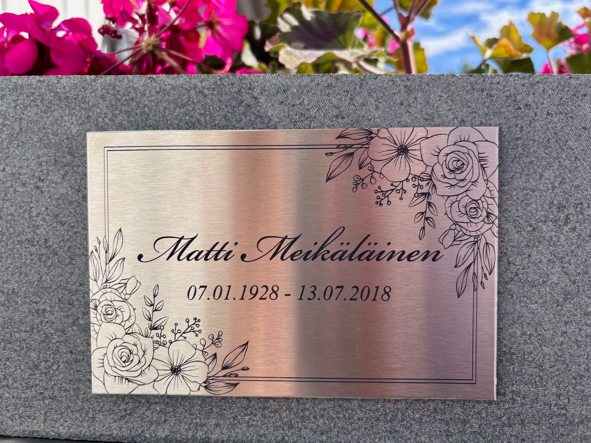 Kaiverrettu muistolaatta, jossa on kukkareunus ja päivämäärät kivipinnalla vaaleanpunaisten kukkien alla