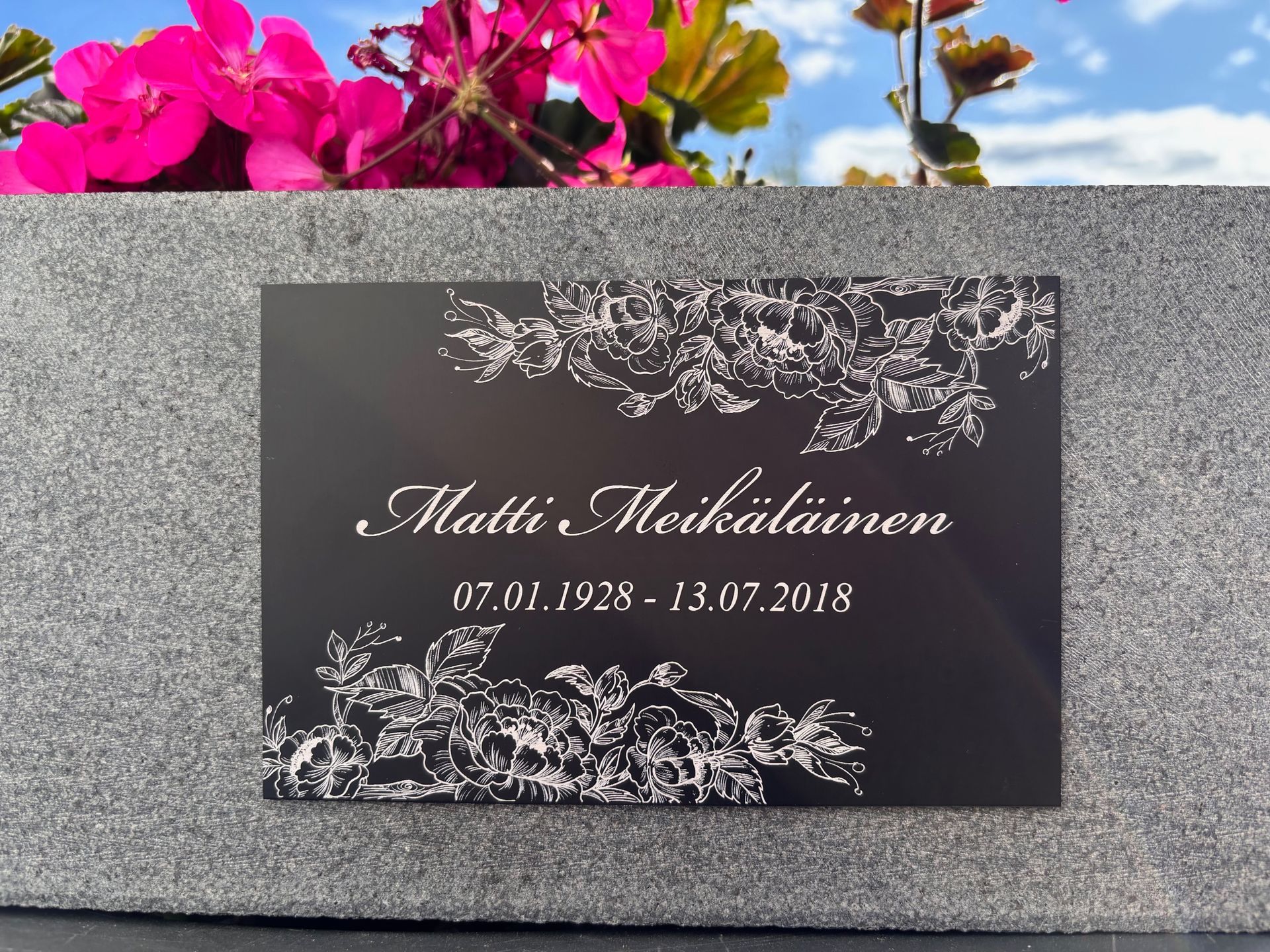 Musta muistolaatta, jossa on valkoinen kukkakuviointi ja nimi Matti Meikäläinen, kiviseinällä.