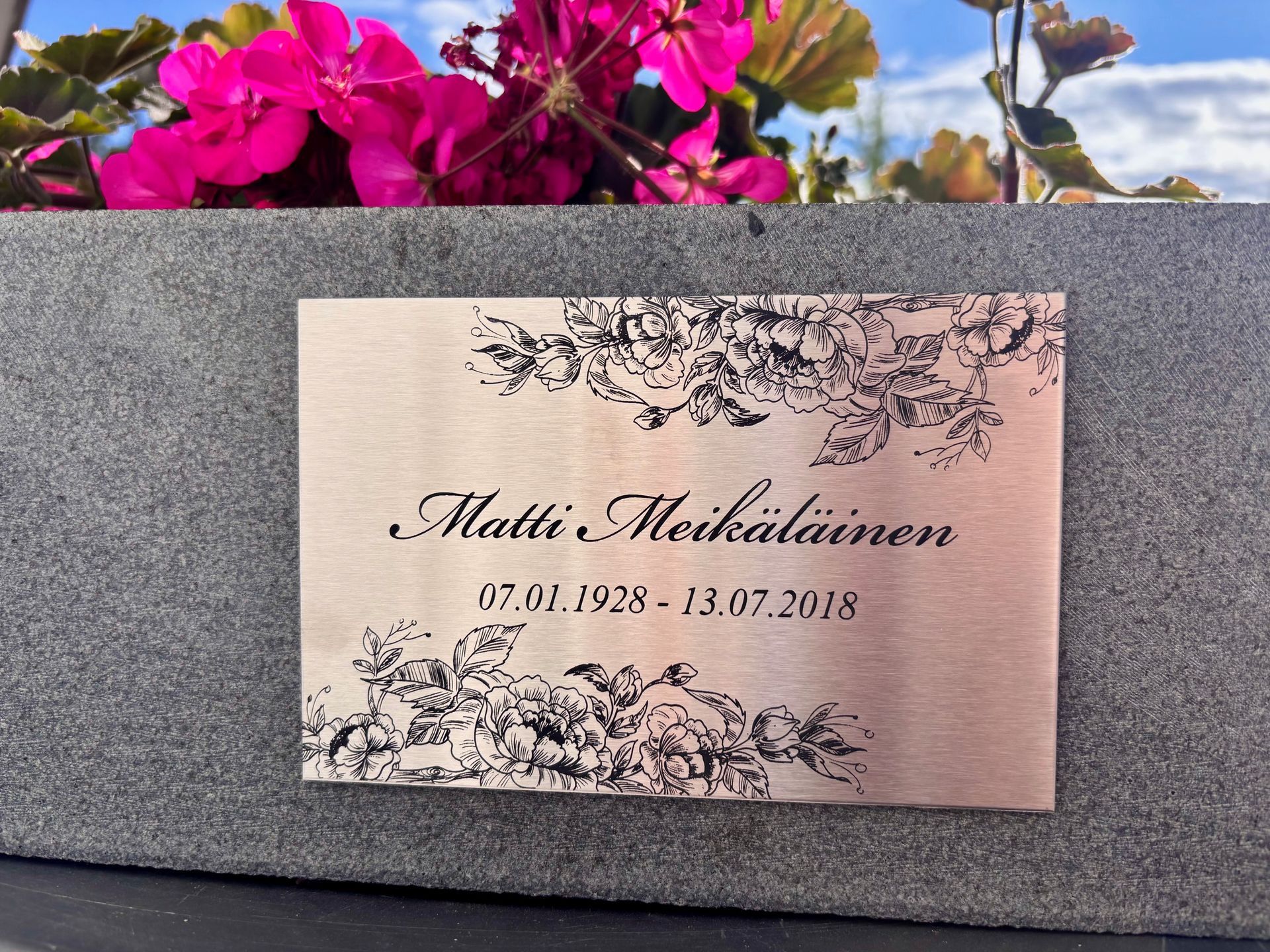 Muistolaatta, jossa on kukkakuvioinen kaiverrus ja nimi harmaalla kivipinnalla vaaleanpunaisten kukkien alla
