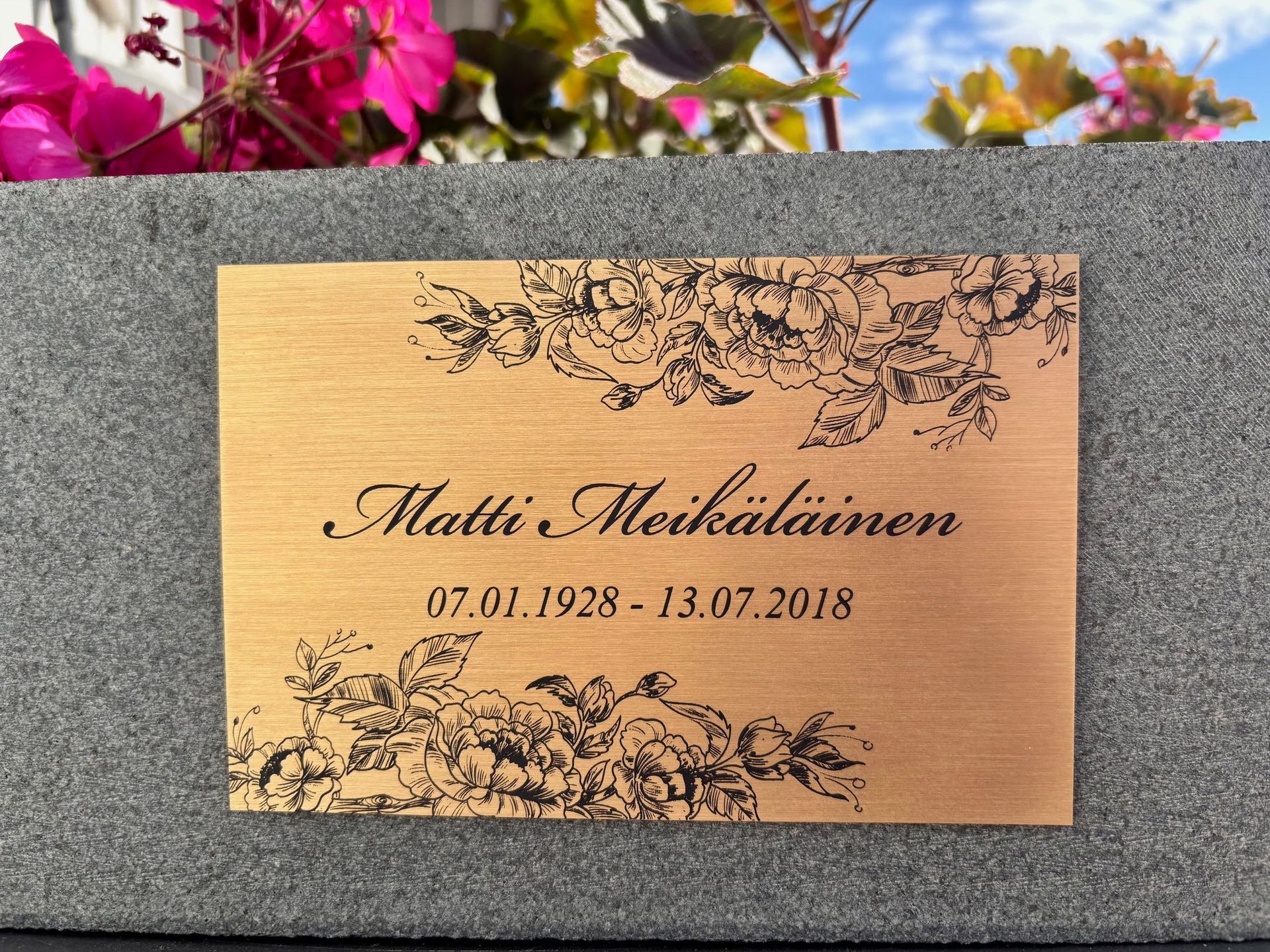 Messinkinen muistolaatta, jossa on kukkakoristeinen reunus. Siihen on kaiverrettu nimi ja päivämäärät.