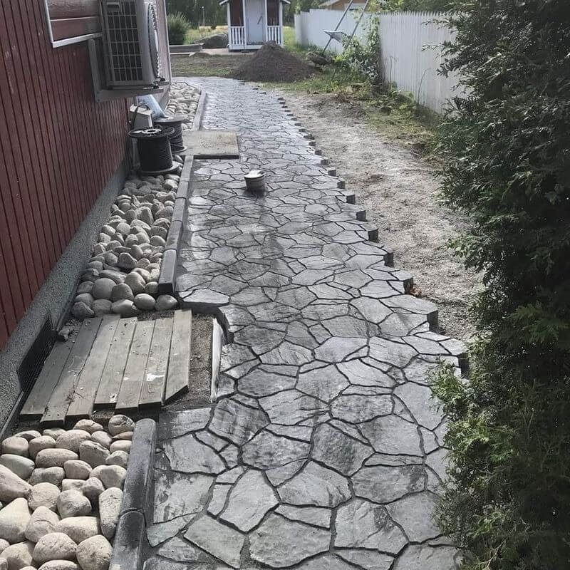 Kapea kivipäällysteinen kävelytie punaisen rakennuksen ja valkoisen aidan vieressä, kivien ja pensaiden reunustamana.