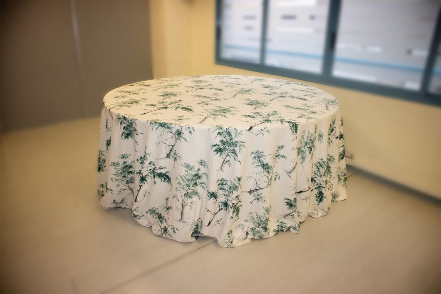 Una mesa redonda cubierta con un mantel color crema con un estampado botánico verde repetitivo en una habitación interior.
