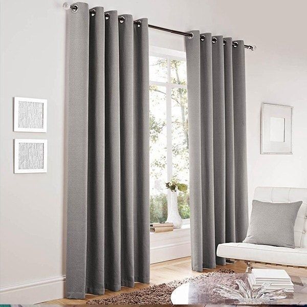Dos cortinas grises con ojales, que llegan hasta el suelo, cuelgan de una barra en una habitación luminosa junto a una silla.