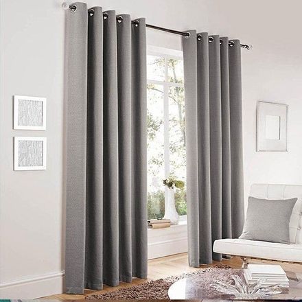 Dos cortinas grises con ojales, que llegan hasta el suelo, cuelgan de una barra en una habitación luminosa junto a una silla.