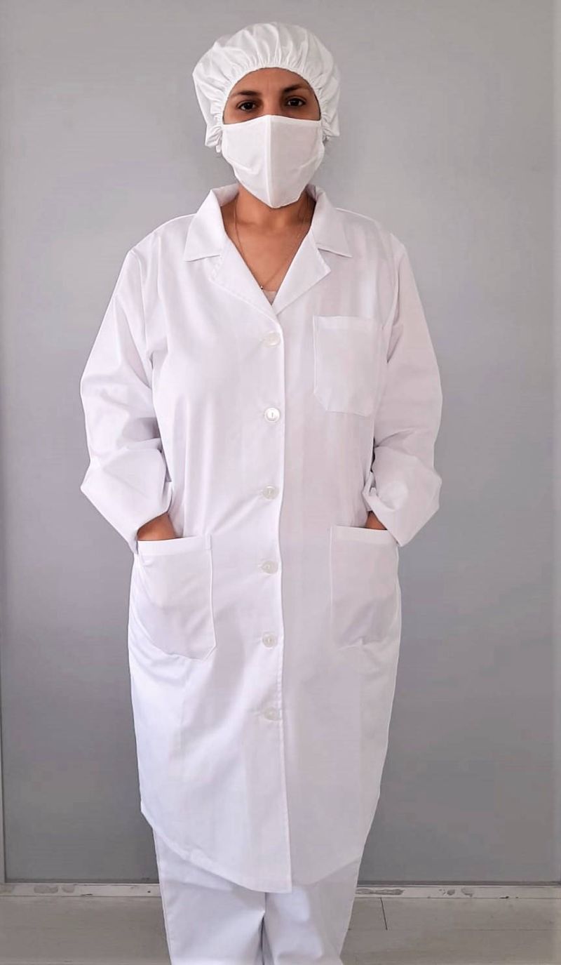Una persona con bata blanca de laboratorio, redecilla para el cabello y mascarilla, de pie contra una pared gris lisa.