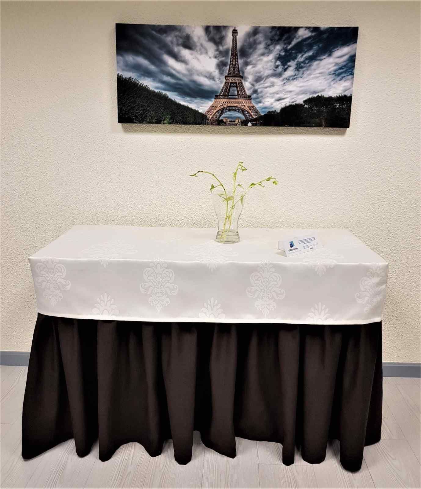 Una mesa rectangular con un mantel blanco estampado y un faldón negro, coronada con un jarrón y un cuadro de la Torre Eiffel.