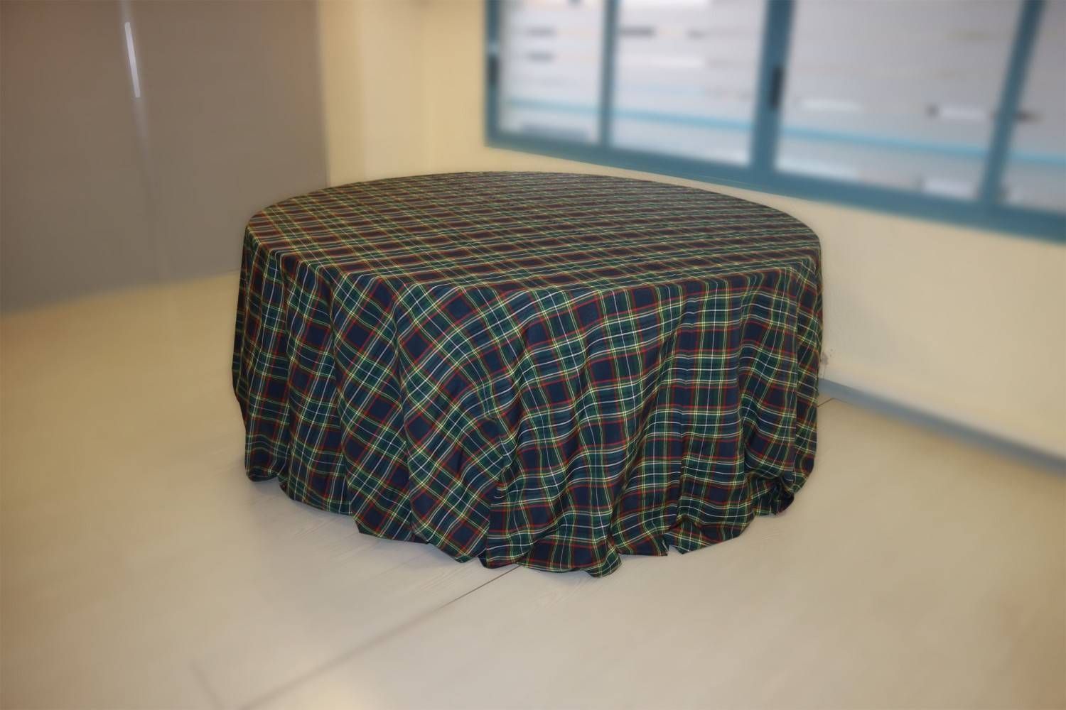 Una mesa redonda cubierta con un mantel a cuadros de color verde oscuro y azul, situada en una habitación luminosa y austera.