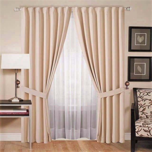 Cortinas beige hasta el suelo con alzapaños, que cuelgan sobre unas cortinas blancas transparentes, junto a una lámpara y un cuadro enmarcado.
