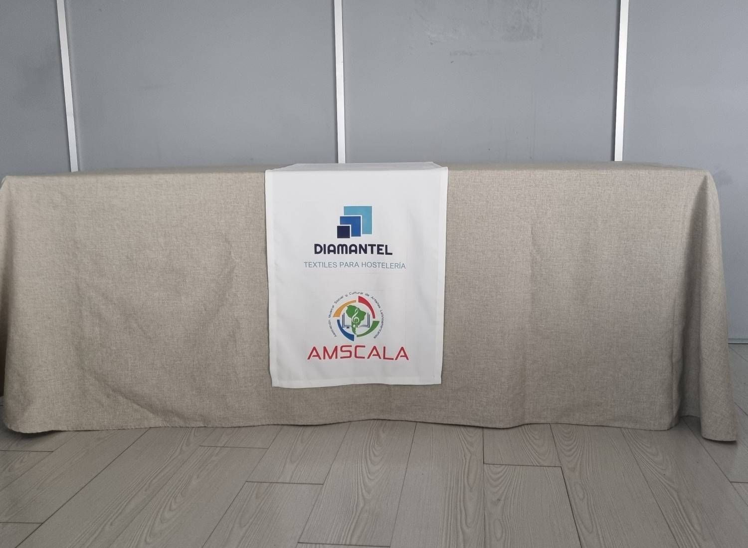 Una mesa cubierta con un mantel de lino de color neutro y un camino de mesa blanco con los logotipos de DamanTel y AMSCALA.