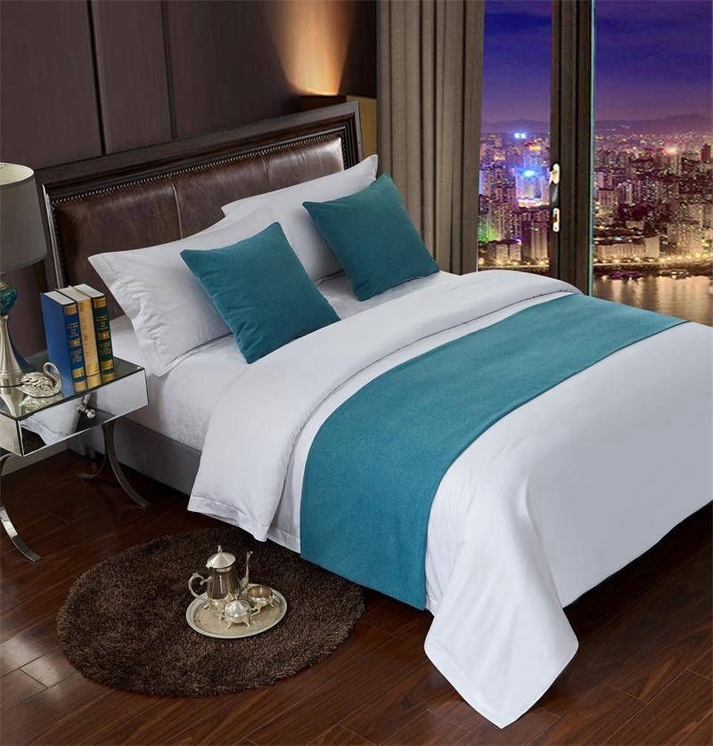 Una cama impecablemente hecha con sábanas blancas, cojines decorativos y un camino de mesa de color turquesa, situada en una habitación moderna con vistas a la ciudad.