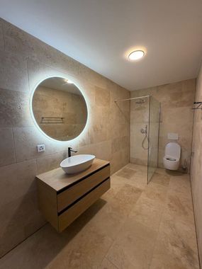 Une salle de bains moderne avec des murs et un sol carrelés couleur beige, comprenant un meuble-lavabo suspendu en bois, une vasque, un miroir rond lumineux et une douche.