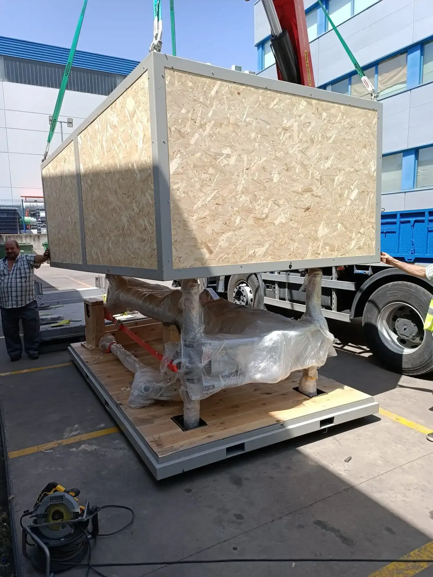 Una grúa levanta una gran caja de madera. Un hombre observa junto a un camión en un muelle de carga.