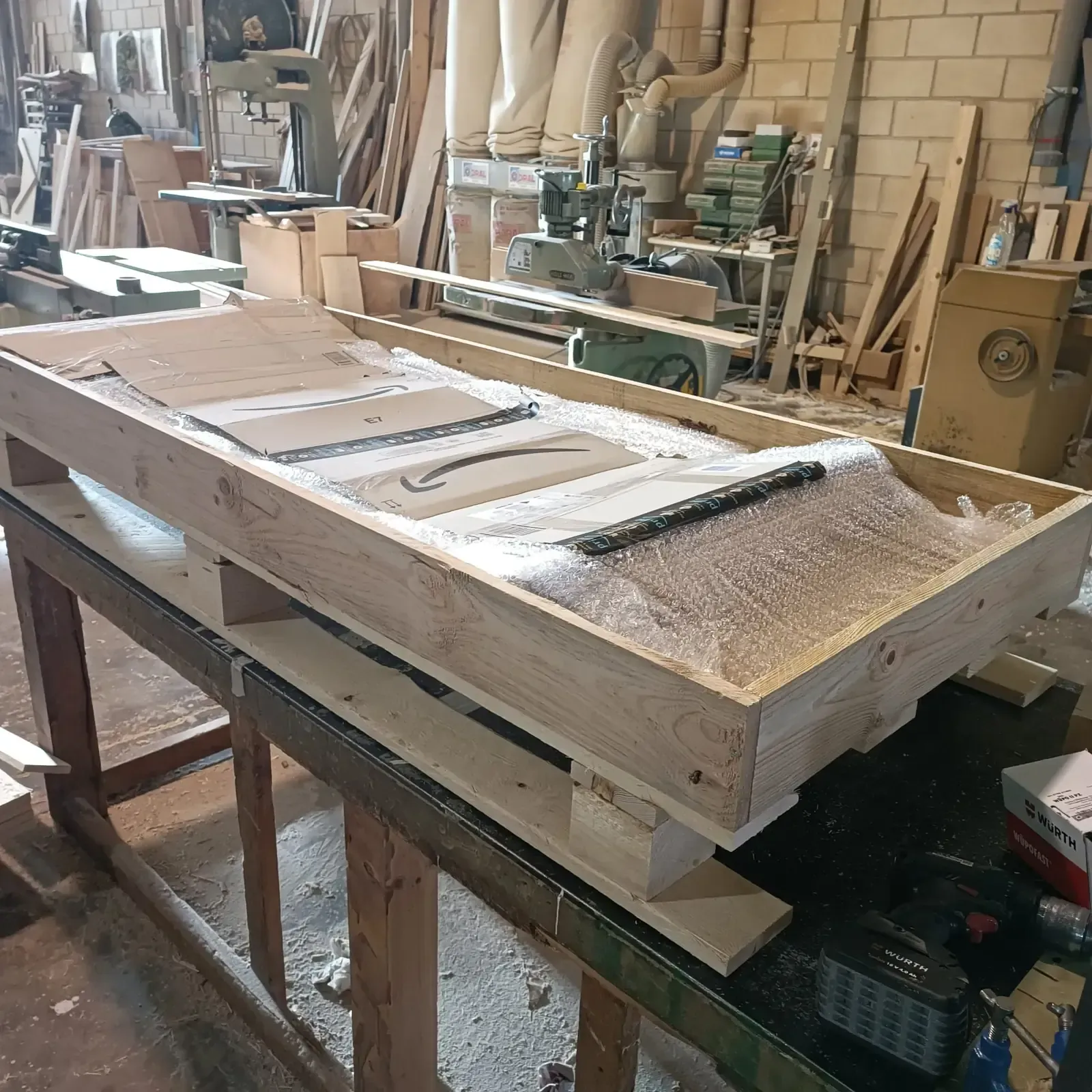 Caja de madera sobre un palé, conteniendo objetos envueltos, dentro de un taller de carpintería.