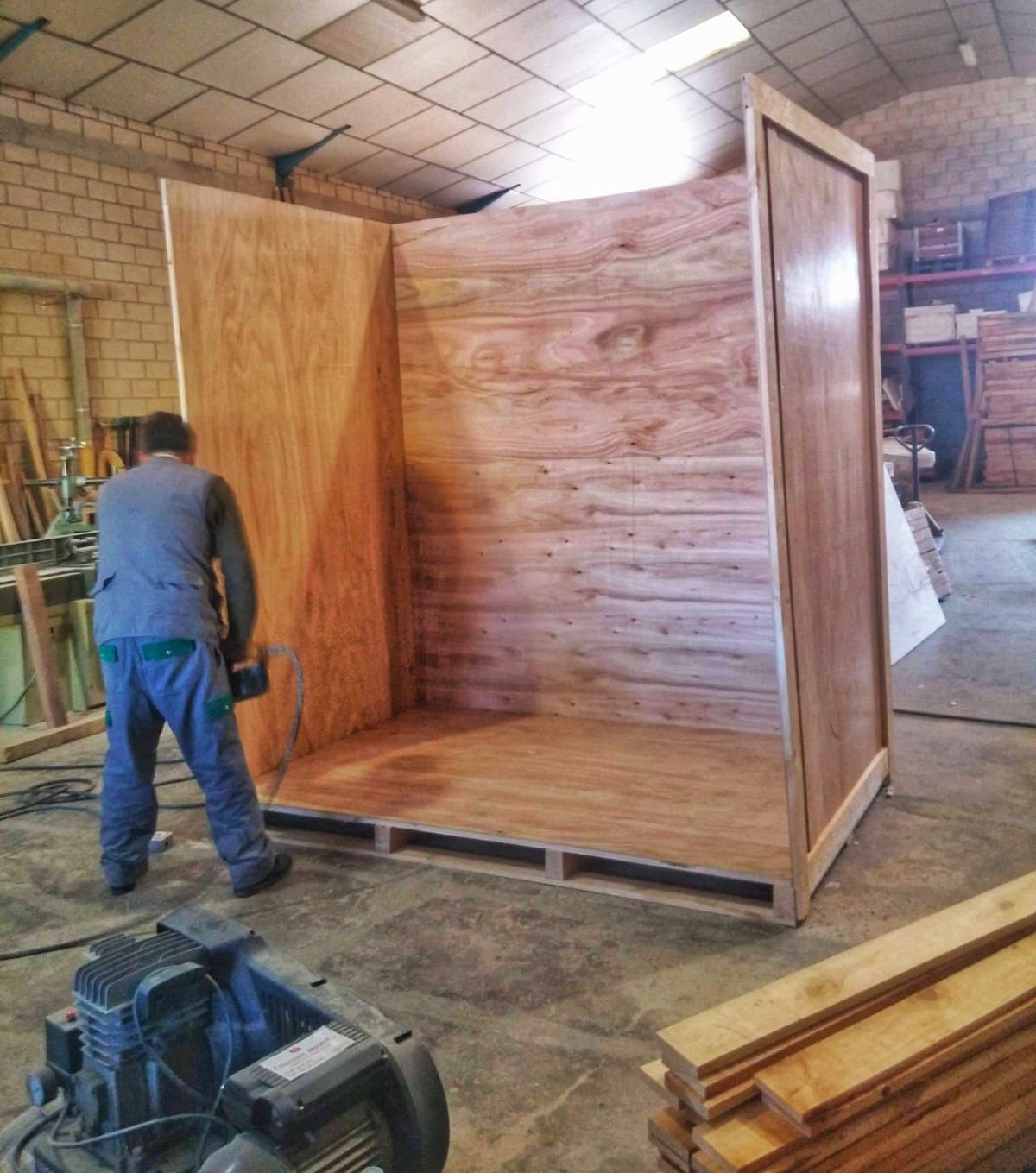 Hombre construyendo una gran caja de madera sobre un palé en un taller. Madera marrón, ropa azul.