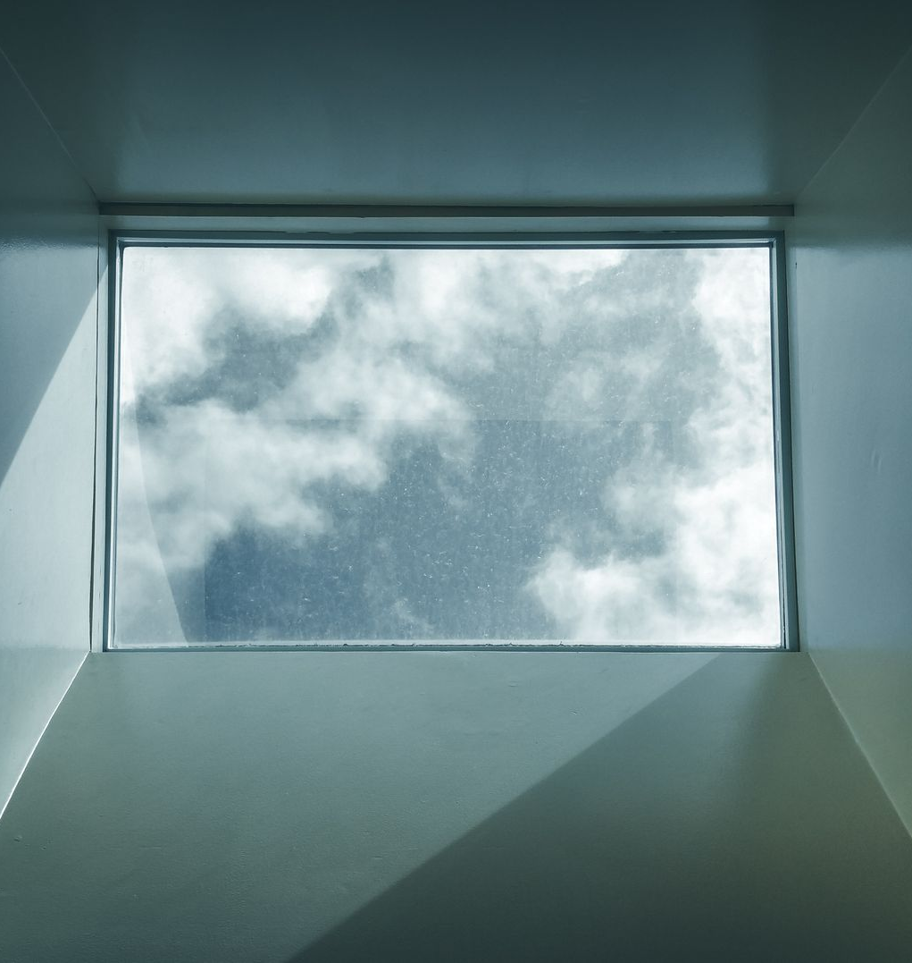 Puits de lumière rectangulaire laissant apparaître un ciel nuageux, où la douce lumière du soleil projette des ombres géométriques sur des murs clairs.
