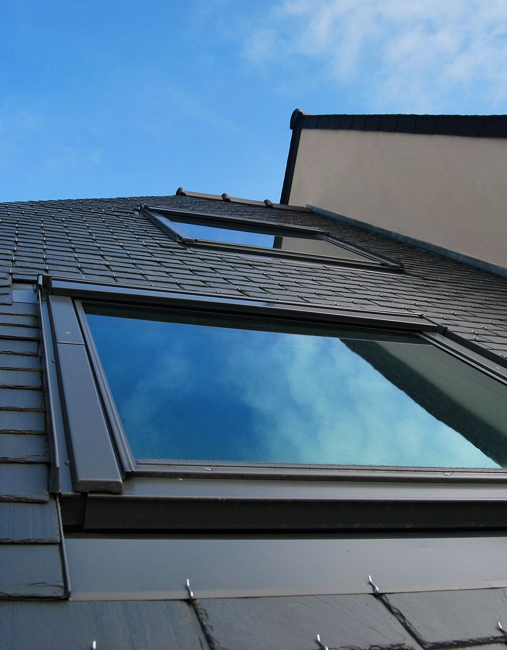 Toit percé d'une fenêtre de toit reflétant un ciel bleu nuageux à côté d'un bâtiment beige