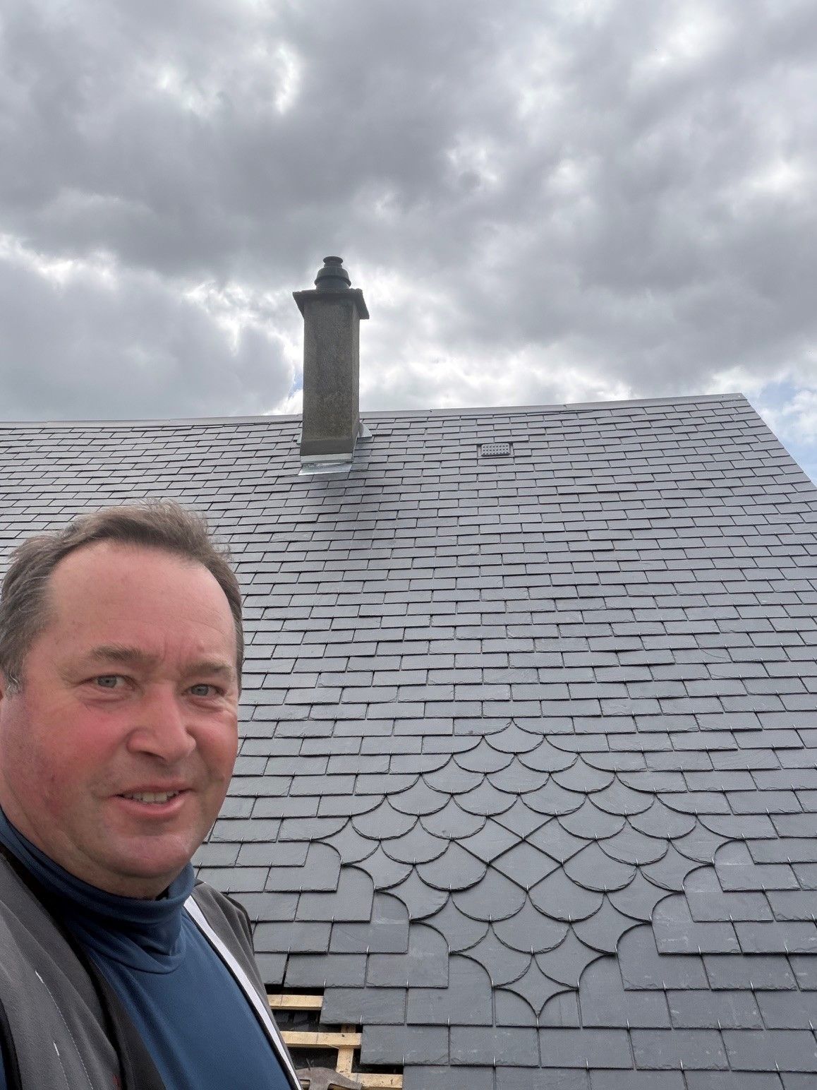 Un homme prend un selfie à côté d'un toit en bardeaux gris et d'une cheminée sous un ciel nuageux