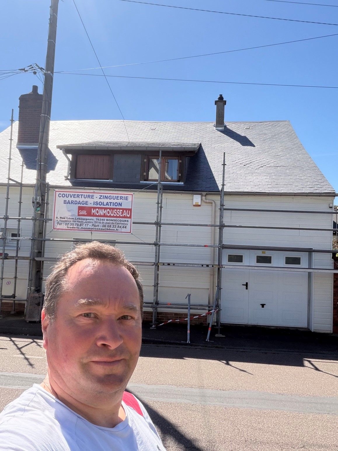 Un homme prend un selfie devant une maison blanche avec un garage et des échafaudages dans une rue ensoleillée.