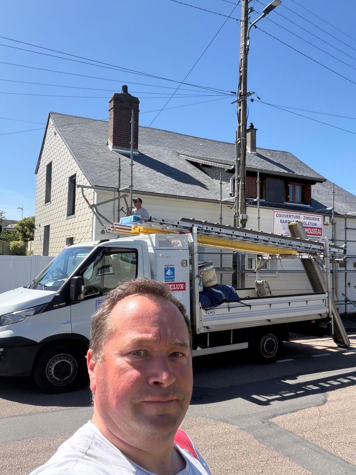 Un homme prend un selfie devant une camionnette blanche et une maison en pierre dans une rue ensoleillée.