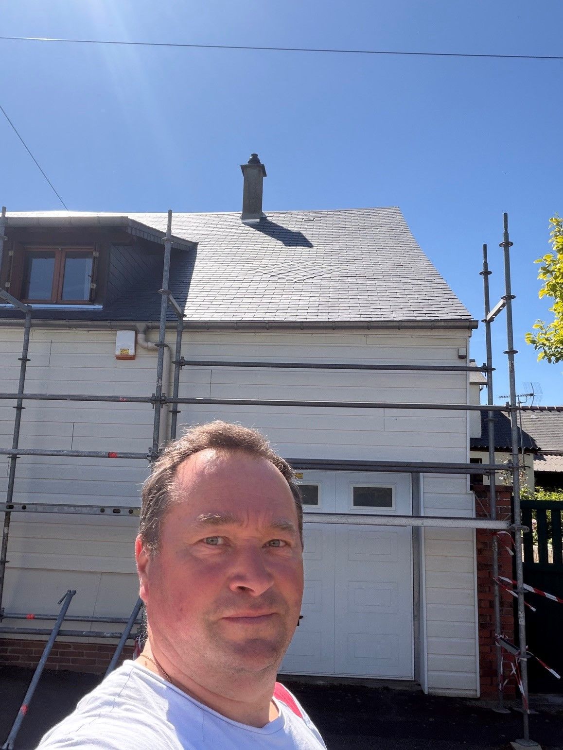 Un homme prend un selfie devant une maison en rénovation par une journée ensoleillée
