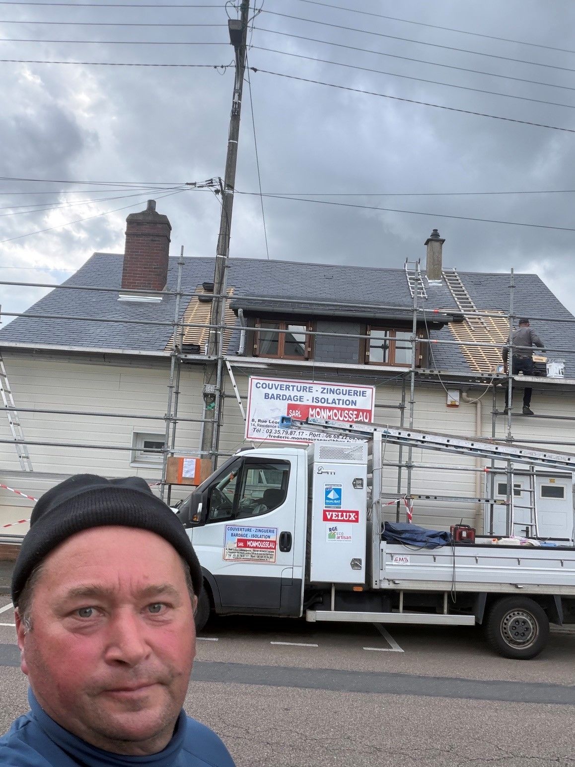 Selfie devant une maison endommagée avec un camion de chantier et des échafaudages sous un ciel nuageux.