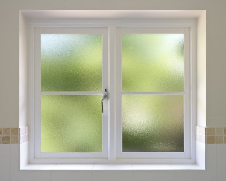 Une fenêtre blanche à double vitrage dépoli, encastrée dans un mur carrelé, laissant entrevoir une vue floue et verdoyante à l'extérieur.