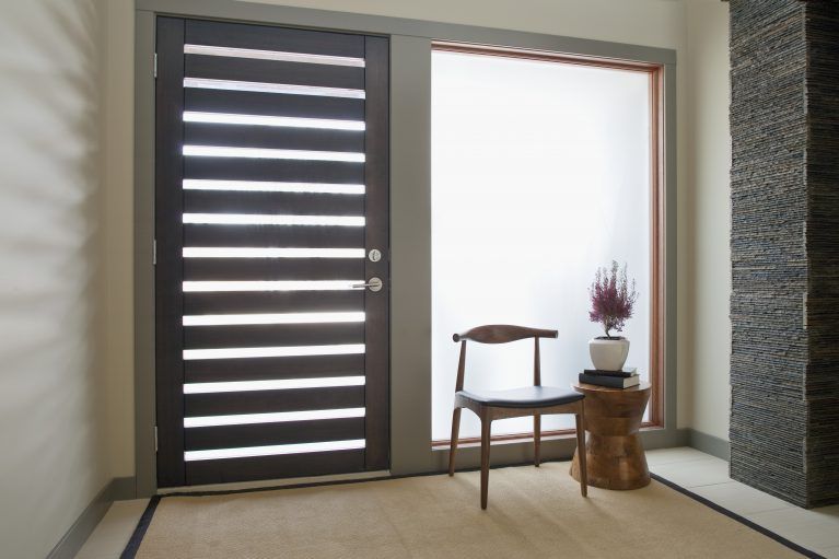 Une porte d'entrée moderne à lattes en bois foncé, jouxtant une fenêtre en verre dépoli, avec une chaise en bois et une petite plante en pot à proximité.