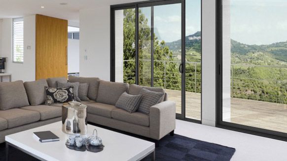 Un salon moderne avec un canapé d'angle gris, une table basse blanche et de grandes portes coulissantes en verre donnant sur un paysage.