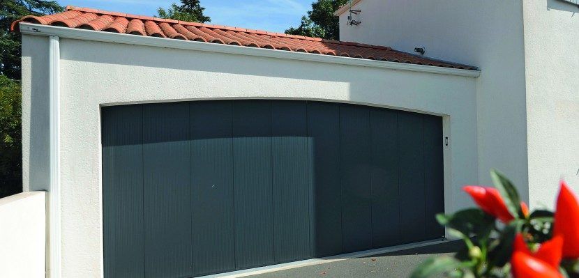 Un garage blanc avec une porte sectionnelle gris foncé et un toit de tuiles en terre cuite, encadré par une plante à fleurs rouges au premier plan.