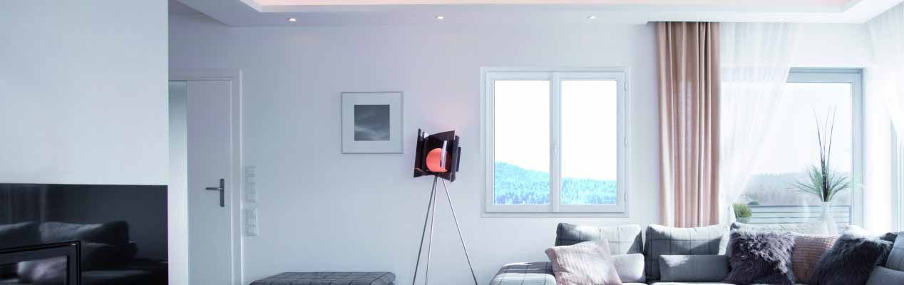 Un salon moderne et lumineux avec des murs clairs, un canapé gris, un lampadaire trépied, un tableau encadré et de grandes fenêtres.