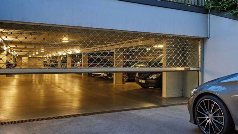 Un portail de sécurité métallique partiellement fermé à l'entrée d'un parking intérieur, une voiture étant visible sur la droite.