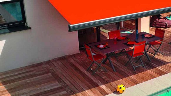 Une table et des chaises à manger sur une terrasse en bois, sous un auvent rétractable orange vif, à côté d'une piscine.