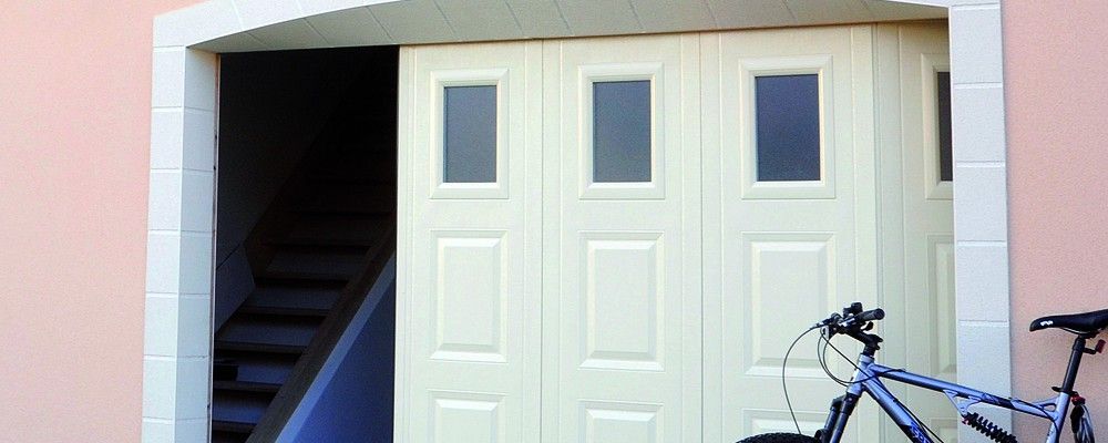 Une porte de garage claire à plusieurs panneaux est légèrement ouverte, révélant un escalier intérieur, avec un vélo à proximité.