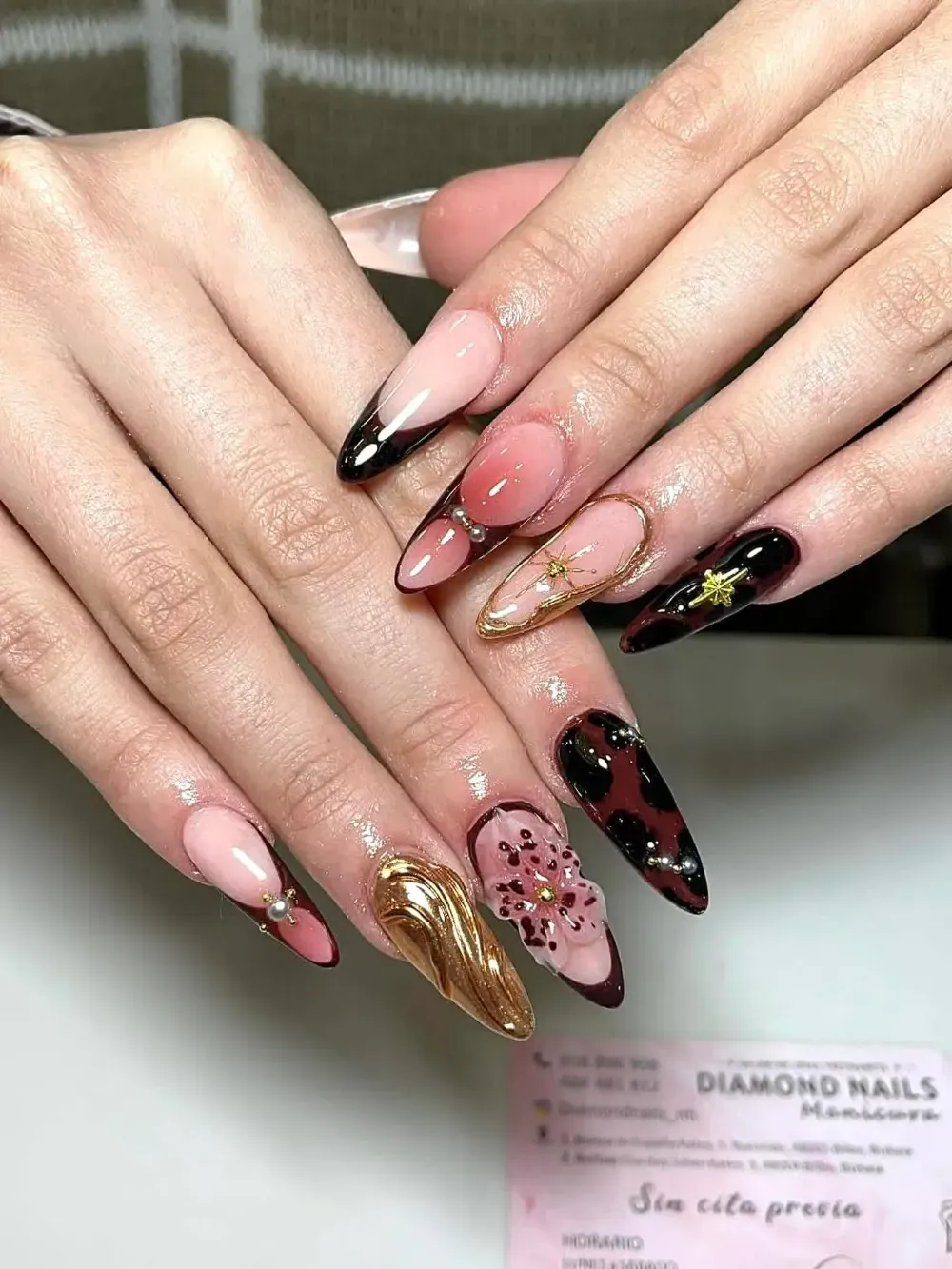 Manos con uñas largas decoradas en rosa, negro y dorado, con diseños de purpurina que muestran un ejemplo de manicura