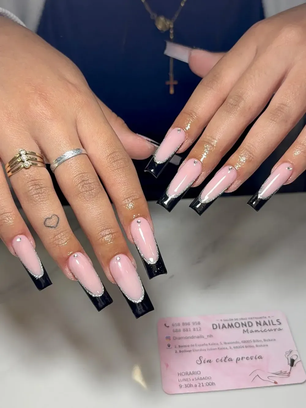 Manos con manicura francesa larga en rosa y negro sobre una tarjeta de un salón de uñas