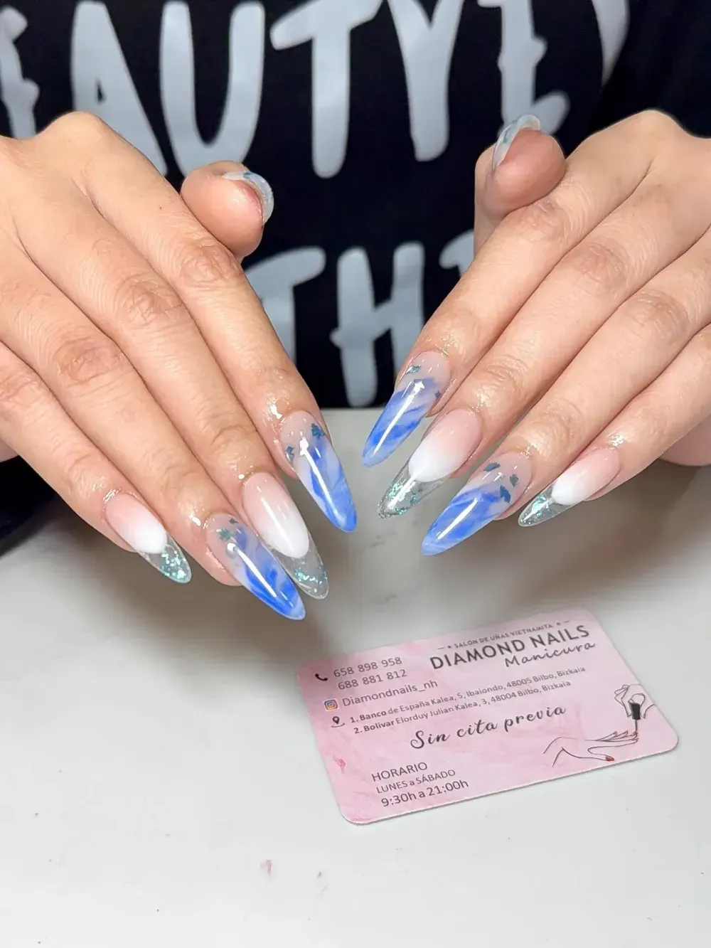 Manos mostrando uñas acrílicas largas con un degradado azul y plateado sobre una tarjeta de un salón de uñas.