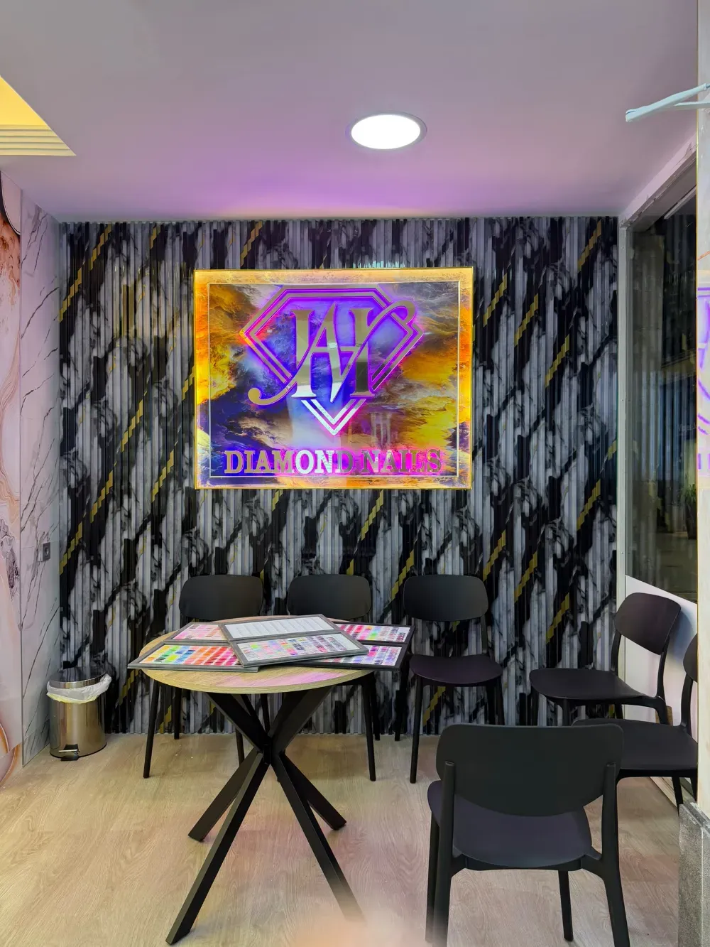 Pequeña zona de espera de una oficina o peluquería, con sillas negras, una mesa auxiliar, papel pintado con efecto mármol y un letrero de neón en la pared.