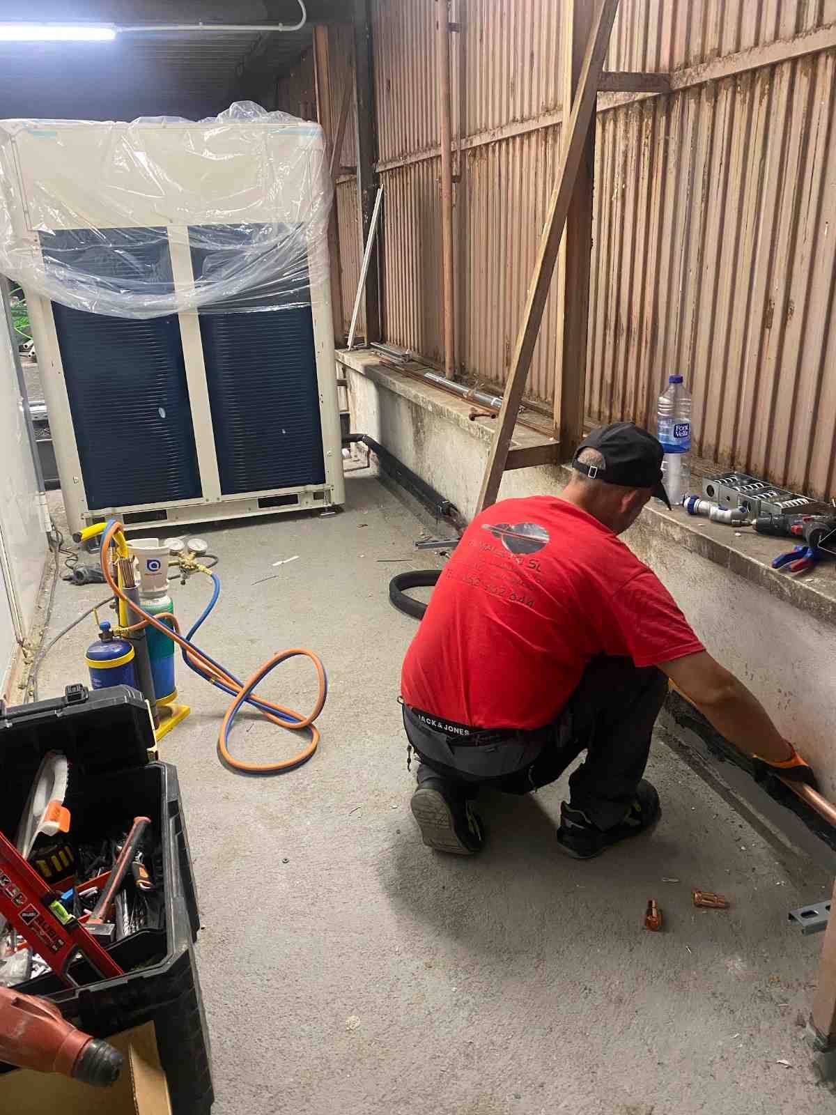 Un trabajador con camisa roja está arrodillado en el suelo, reparando una pared en un taller con herramientas y cables cerca.