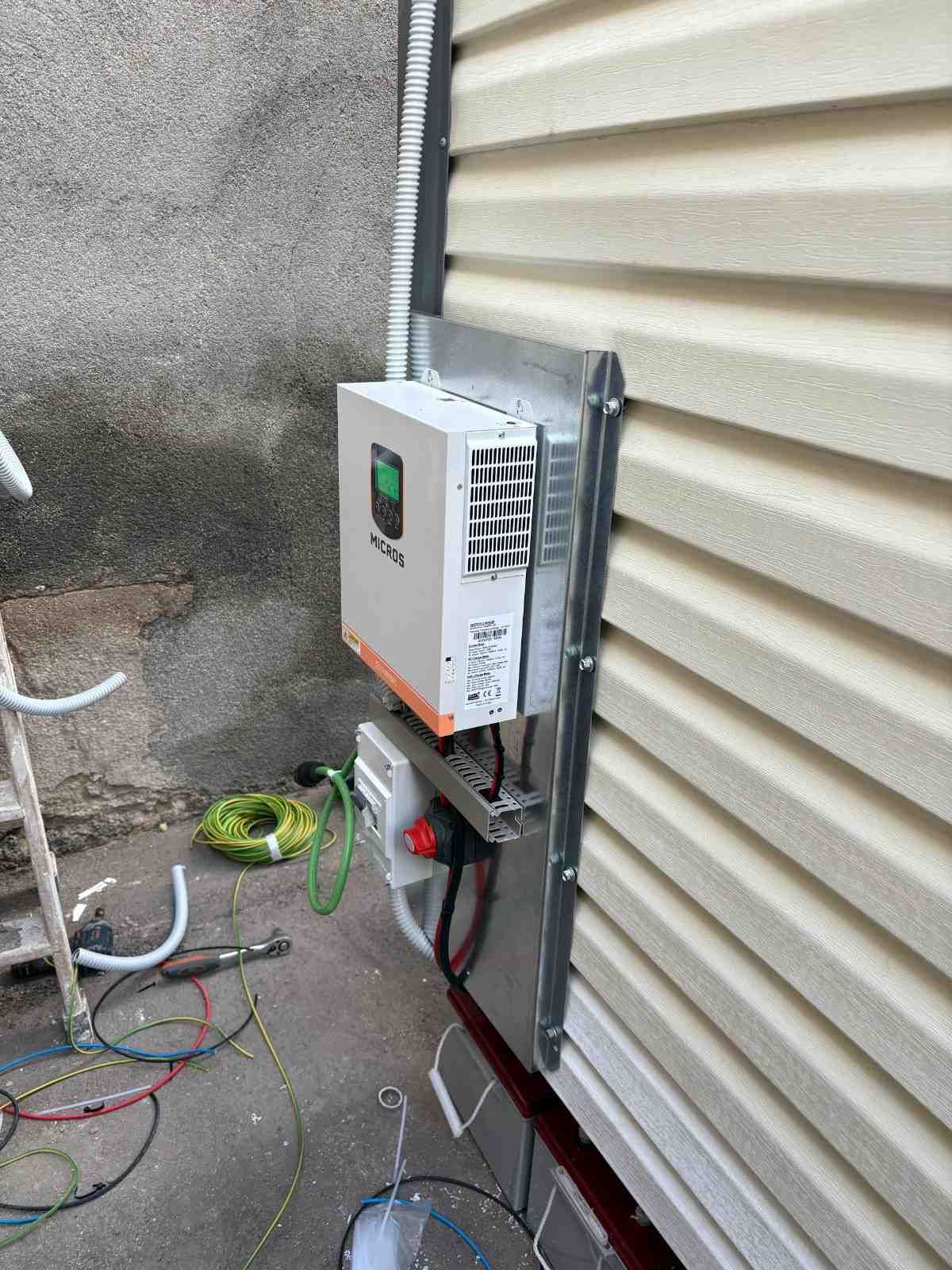 Caja eléctrica exterior montada en la pared con cables y alambres junto a un revestimiento beige y un patio de concreto.