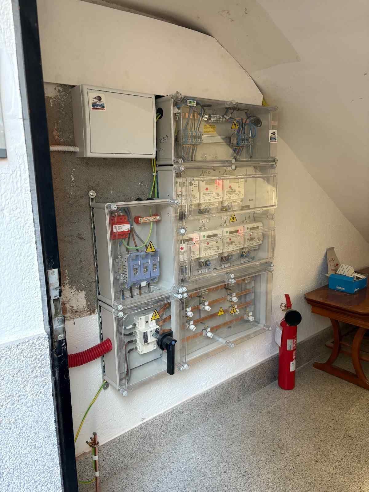 Panel eléctrico y armario de cableado en la pared de una escalera, con un extintor rojo al lado.