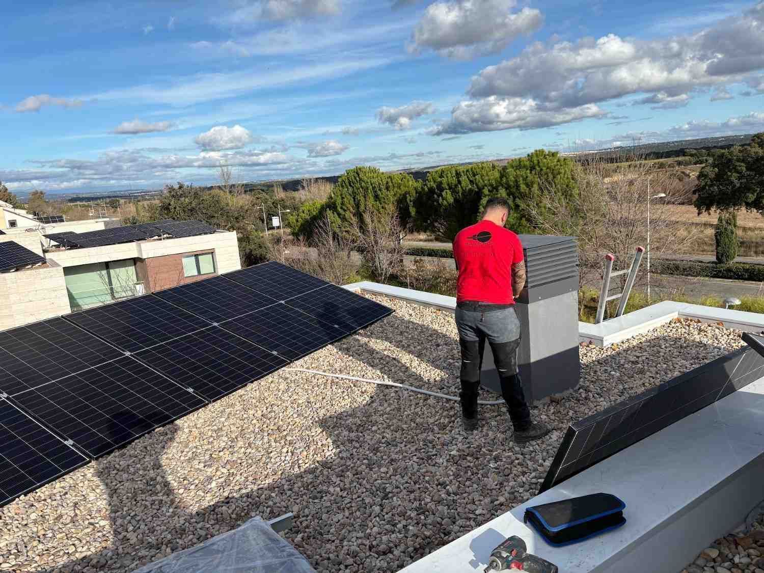Una persona con camisa roja está de pie sobre un tejado de grava junto a unos paneles solares bajo un cielo nublado.