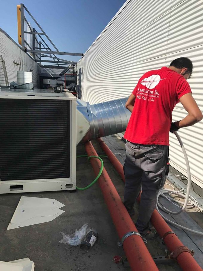 Un trabajador vestido de rojo instala conductos metálicos junto a una unidad de climatización en la azotea.