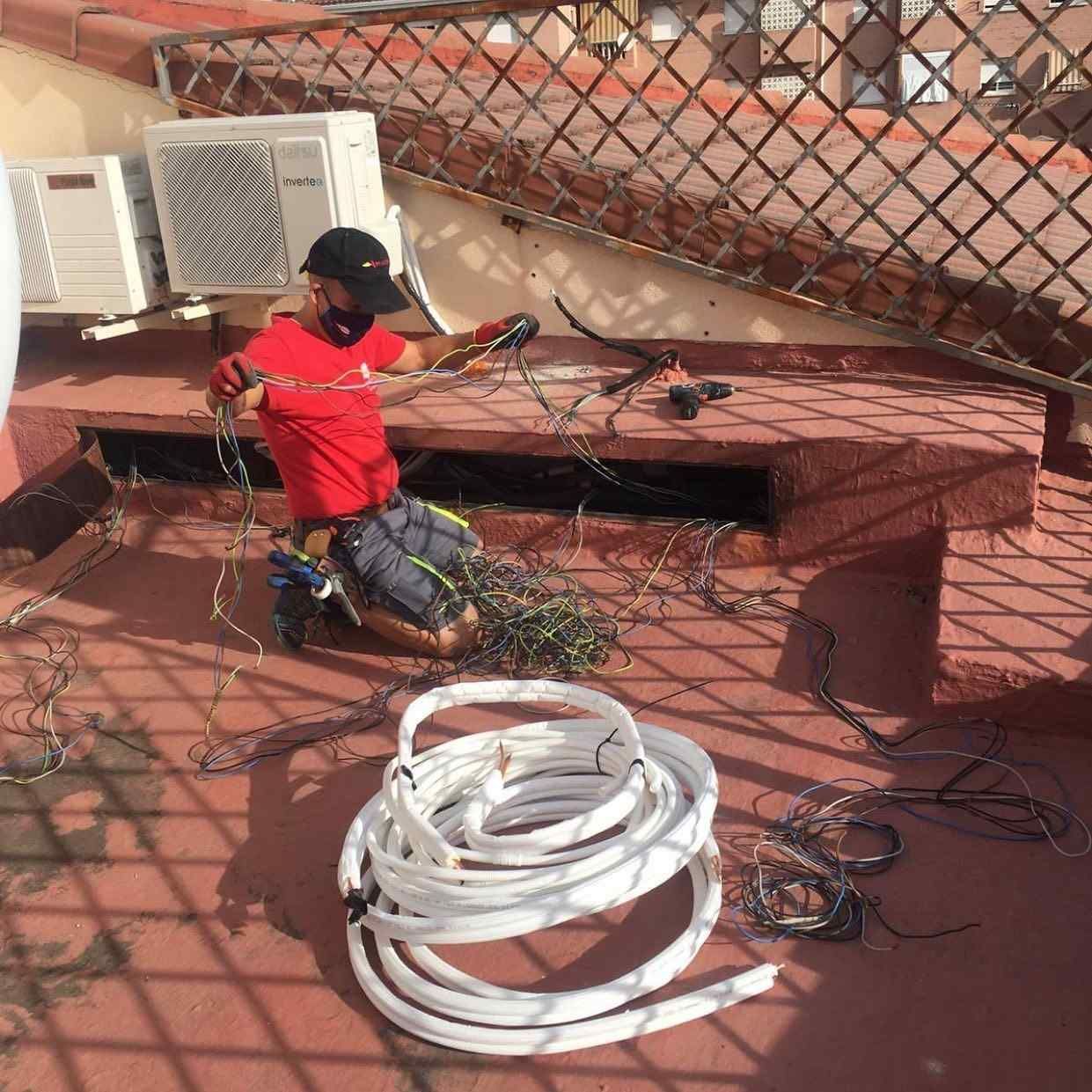 Un trabajador con una camisa roja está arrodillado en una azotea, manipulando cables junto a un rollo de tubería blanca.
