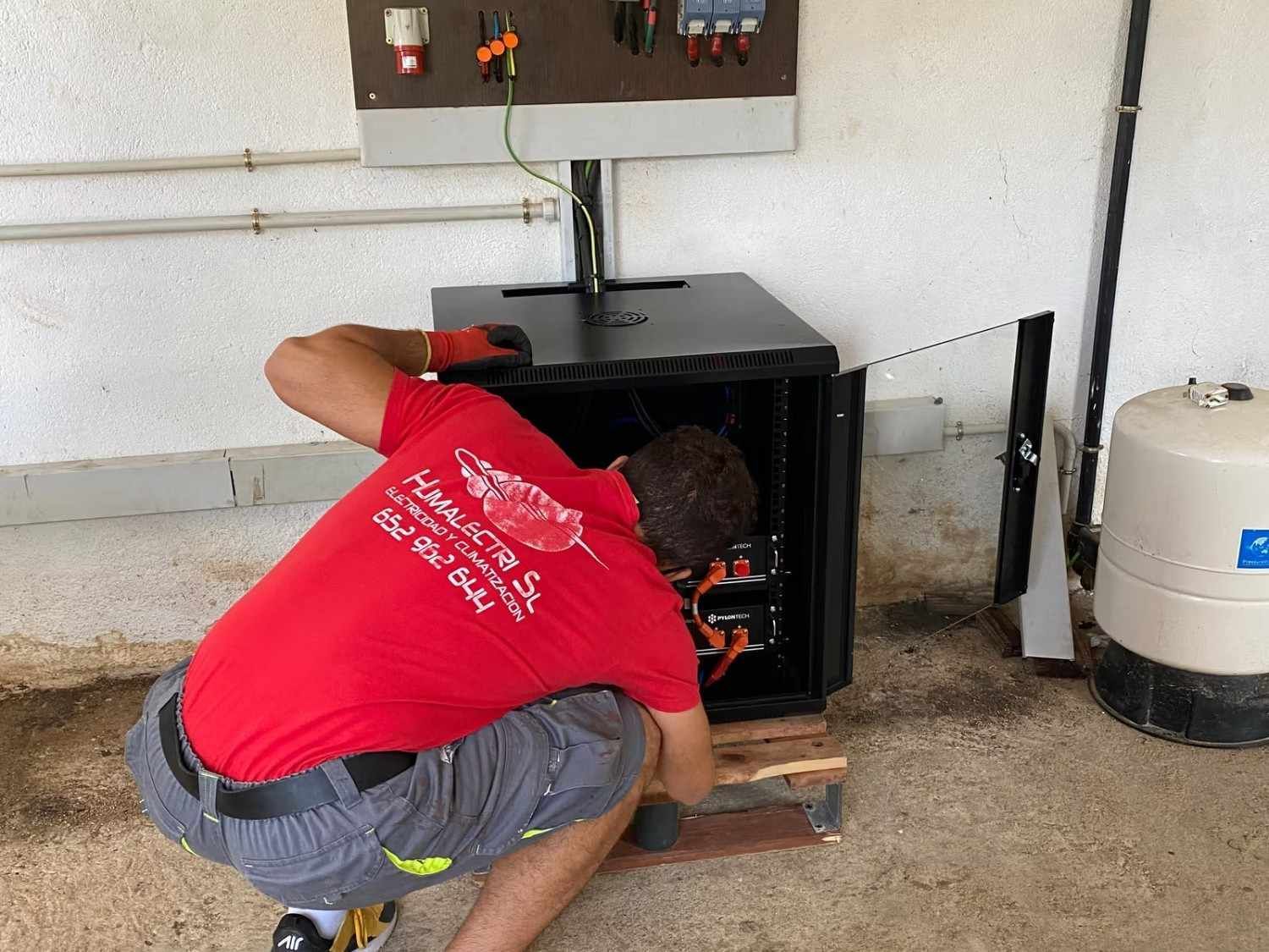 Una persona con una camisa roja está agachada junto a un horno o estufa negra, trabajando en su interior en un garaje.