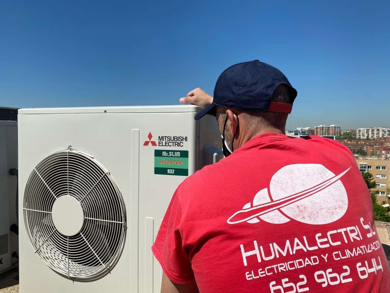 Un técnico con camisa roja inspecciona una unidad eléctrica Mitsubishi en la azotea bajo el cielo azul.