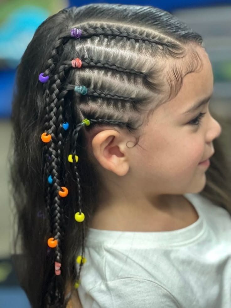 Niña pequeña con el pelo trenzado y cuentas de colores, vista de perfil en el interior de un edificio.