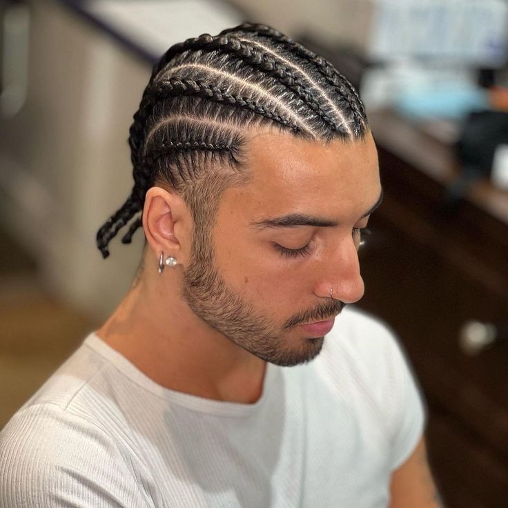 Hombre con trenzas africanas y barba recortada, mirando hacia abajo, vestido con una camisa blanca.