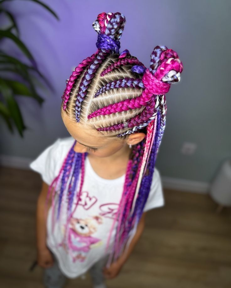 Niña con el pelo trenzado de color rosa y morado, vestida con una camiseta blanca, de pie en el interior de una vivienda.